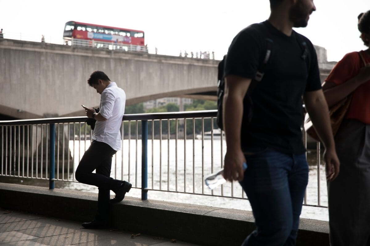 A man leaning against a fence along the Thames River looking at mobile phone